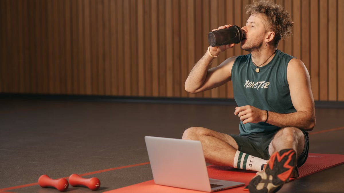 Man in gym drinking water during workout on exercise mat with laptop.
